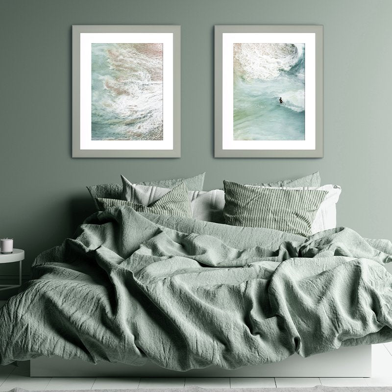 Zwei Pastellrahmen hängen in einem modernen Schalfzimmer über einem Bett an einer Pastellgrünen Wand mit Kunstdrucken vom Meer. 