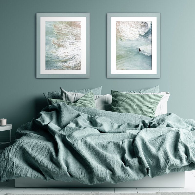 Zwei Pastellrahmen hängen in einem modernen Schalfzimmer über einem Bett an einer Pastelltürkisen Wand mit Kunstdrucken vom Meer. 