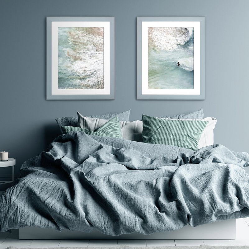 Zwei Pastellrahmen hängen in einem modernen Schalfzimmer über einem Bett an einer Pastellblauen Wand mit Kunstdrucken vom Meer. 